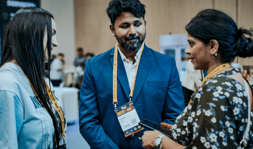 Three individuals engage in conversation at an event, with one person holding a smartphone and wearing a name badge.