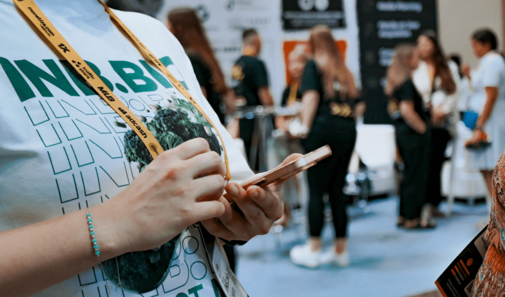 A person in a printed t-shirt checks their phone, with an exhibition backdrop featuring attendees in casual attire.