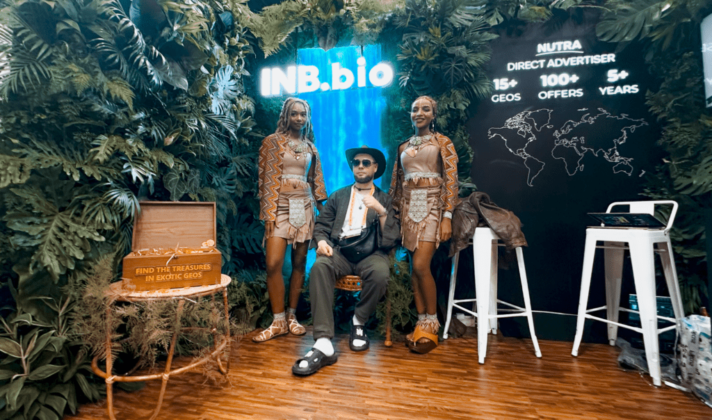 A person sits between two individuals in colorful outfits against a lush green backdrop, featuring a decorative treasure chest nearby.