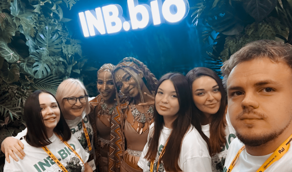 A group of people in matching INB.bio t-shirts smiling in front of a vibrant green foliage backdrop and a glowing sign.