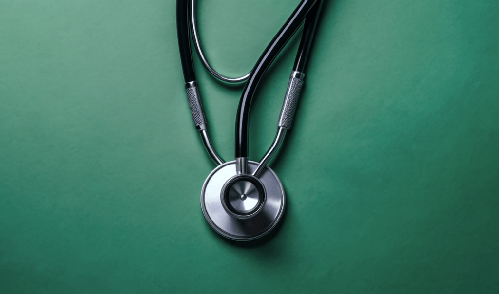 A silver stethoscope rests on a green background, showcasing its circular chest piece and black tubing with texturized grips.
