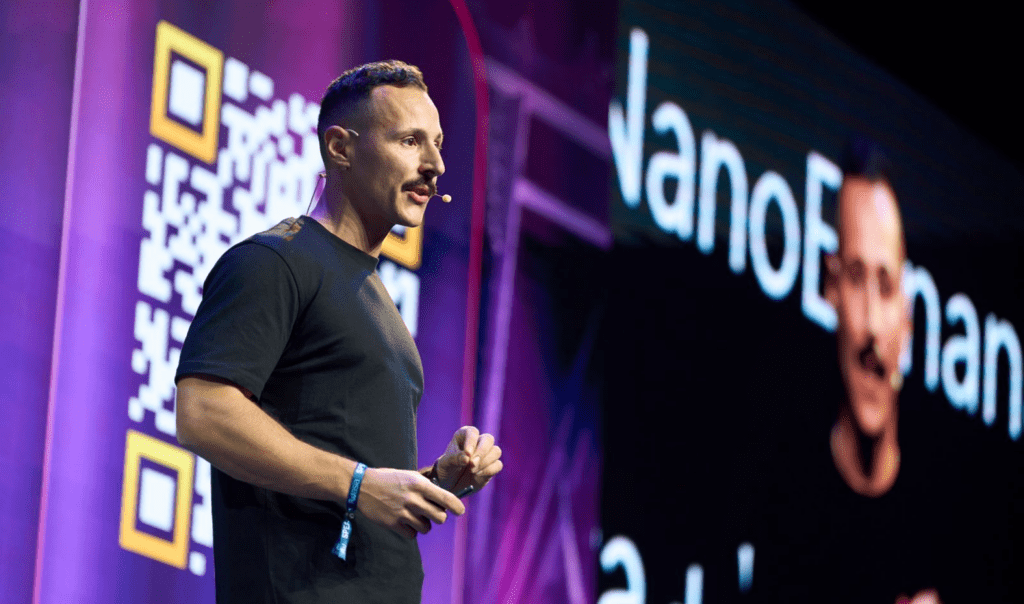 A speaker gestures on stage, with a QR code and a digital screen displaying text in the background. The scene has vibrant lighting.