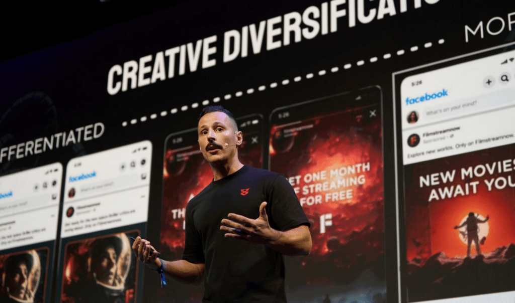 A speaker gestures expressively in front of a presentation displaying mobile app screenshots and marketing promotions.