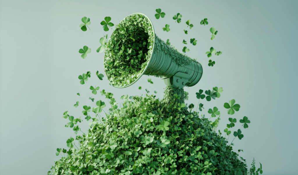 A megaphone covered in green clovers sits atop a mound of foliage, with additional clovers scattering in the air against a soft background.