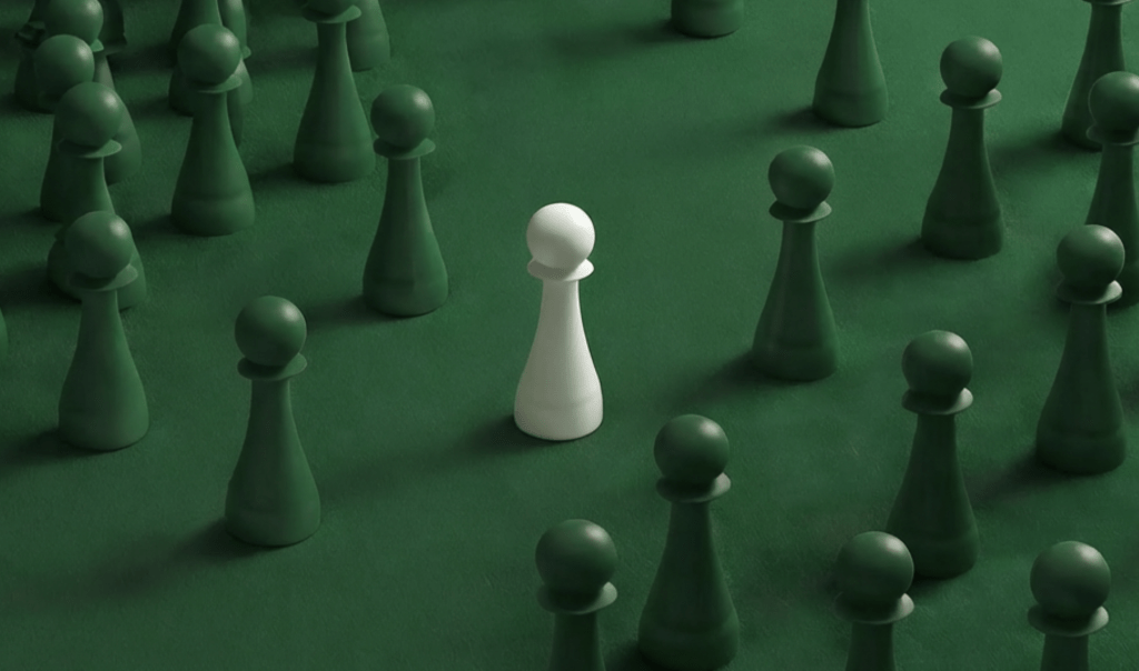 A lone white pawn stands amidst a cluster of green pawns on a green felt surface, symbolizing individuality in a crowd.