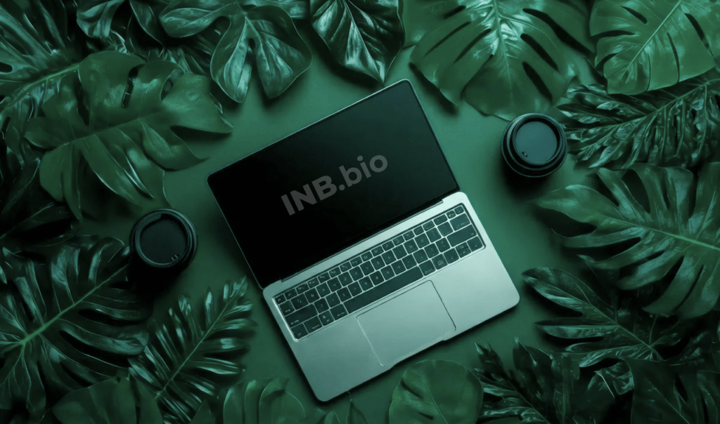 A laptop with the text "INB.bio" sits among lush green leaves, accompanied by two coffee cups, on a dark green background.
